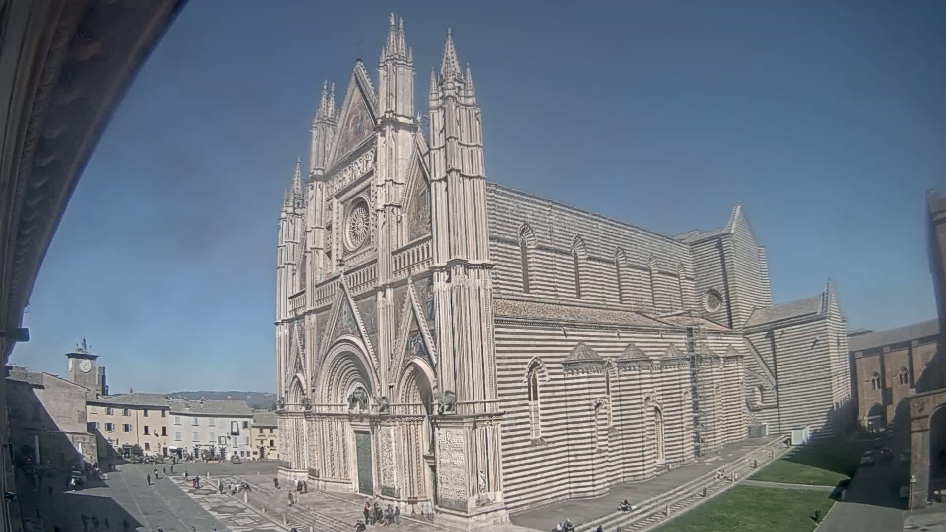 A large, striped cathedral stands in a sunny plaza, with other buildings and a few people visible in the background.