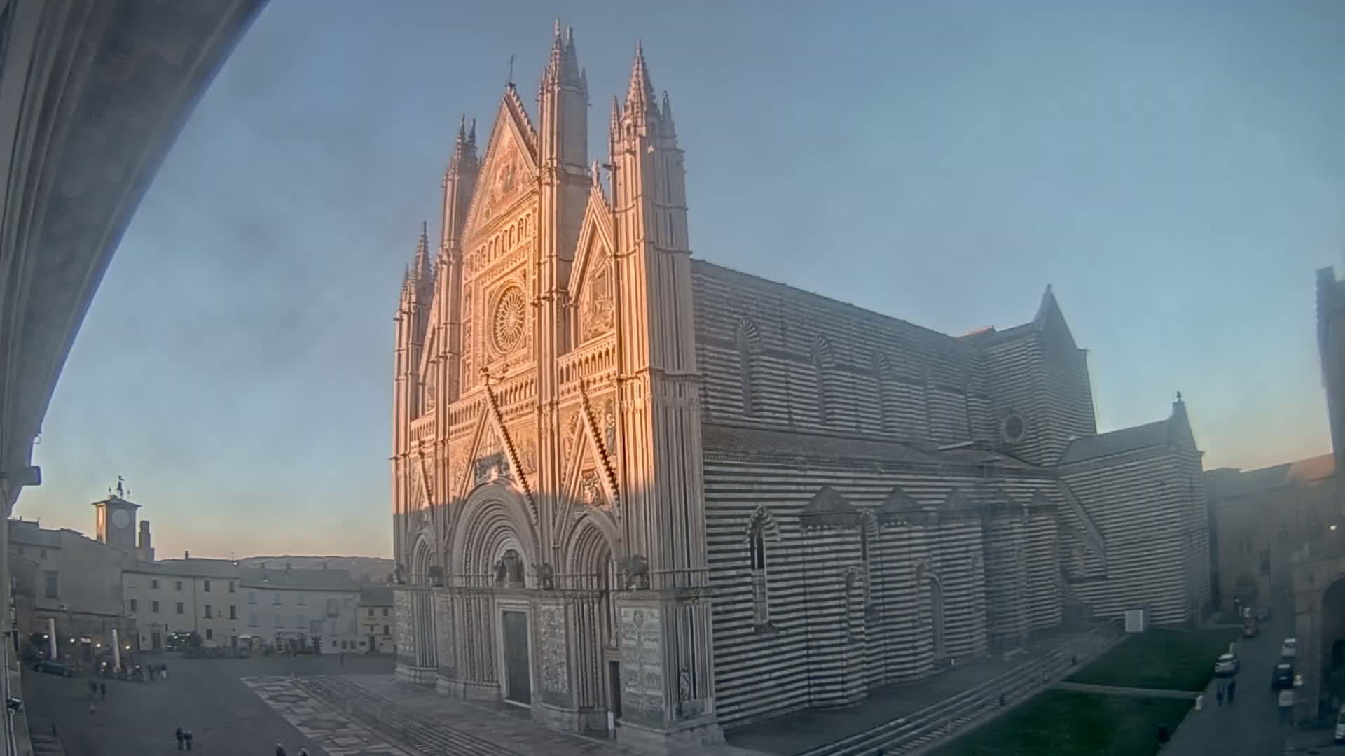A large, striped cathedral stands in a town square under a clear, pale blue sky at sunset.
