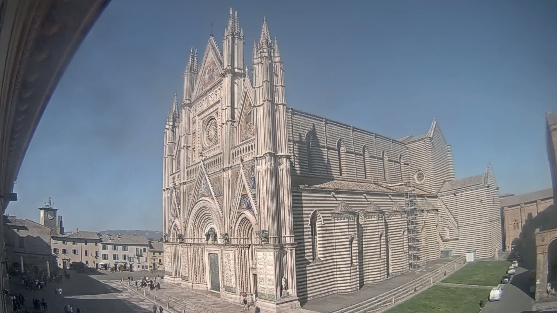 A large, striped cathedral stands in a sunny plaza with a few people and vehicles visible in the distance.