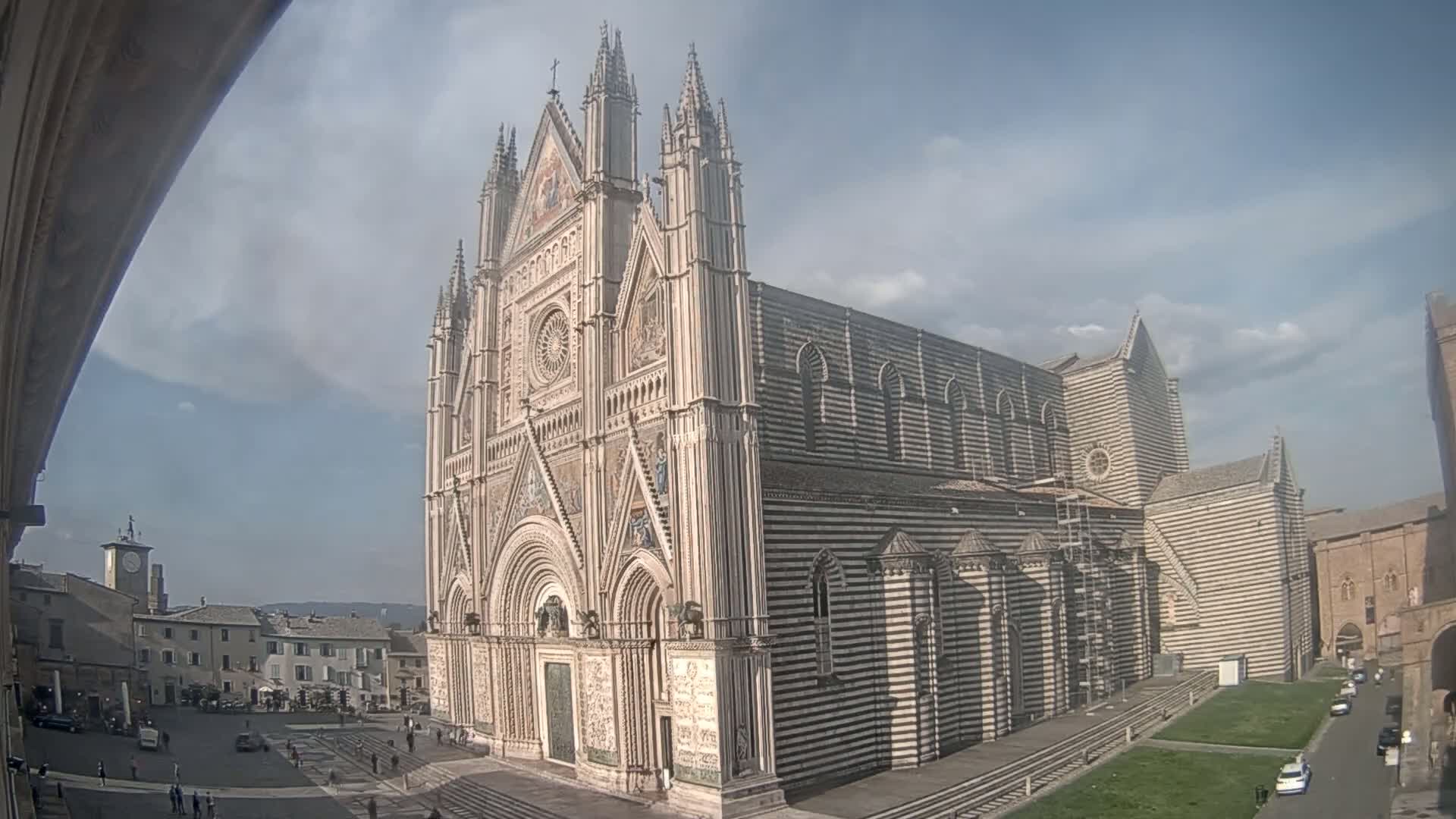 A large, ornate cathedral with striped exterior walls stands in a town square under a partly cloudy sky.