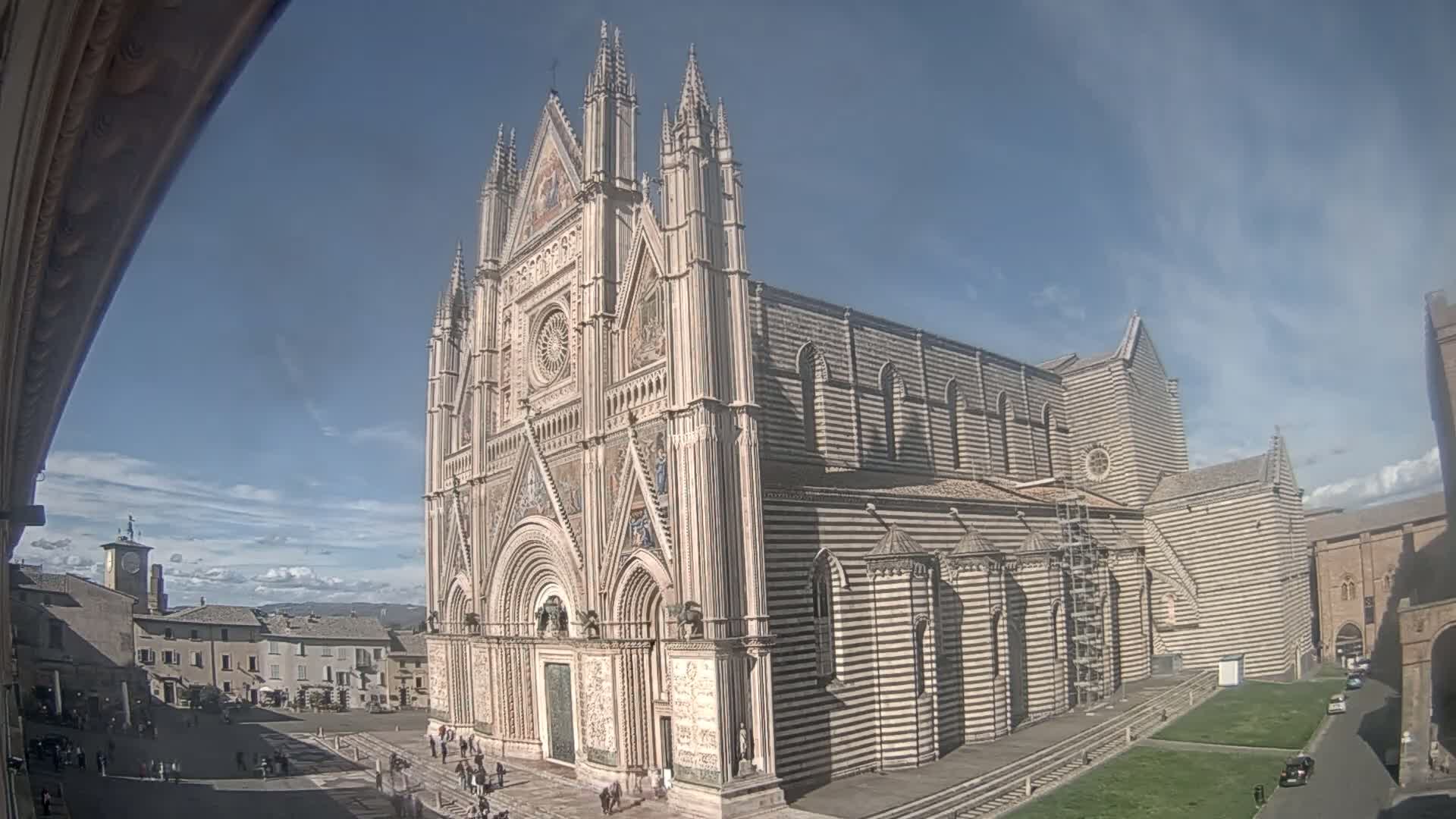 A large ornate cathedral with striped stonework stands in a sunny plaza, under a mostly clear blue sky with a few clouds.
