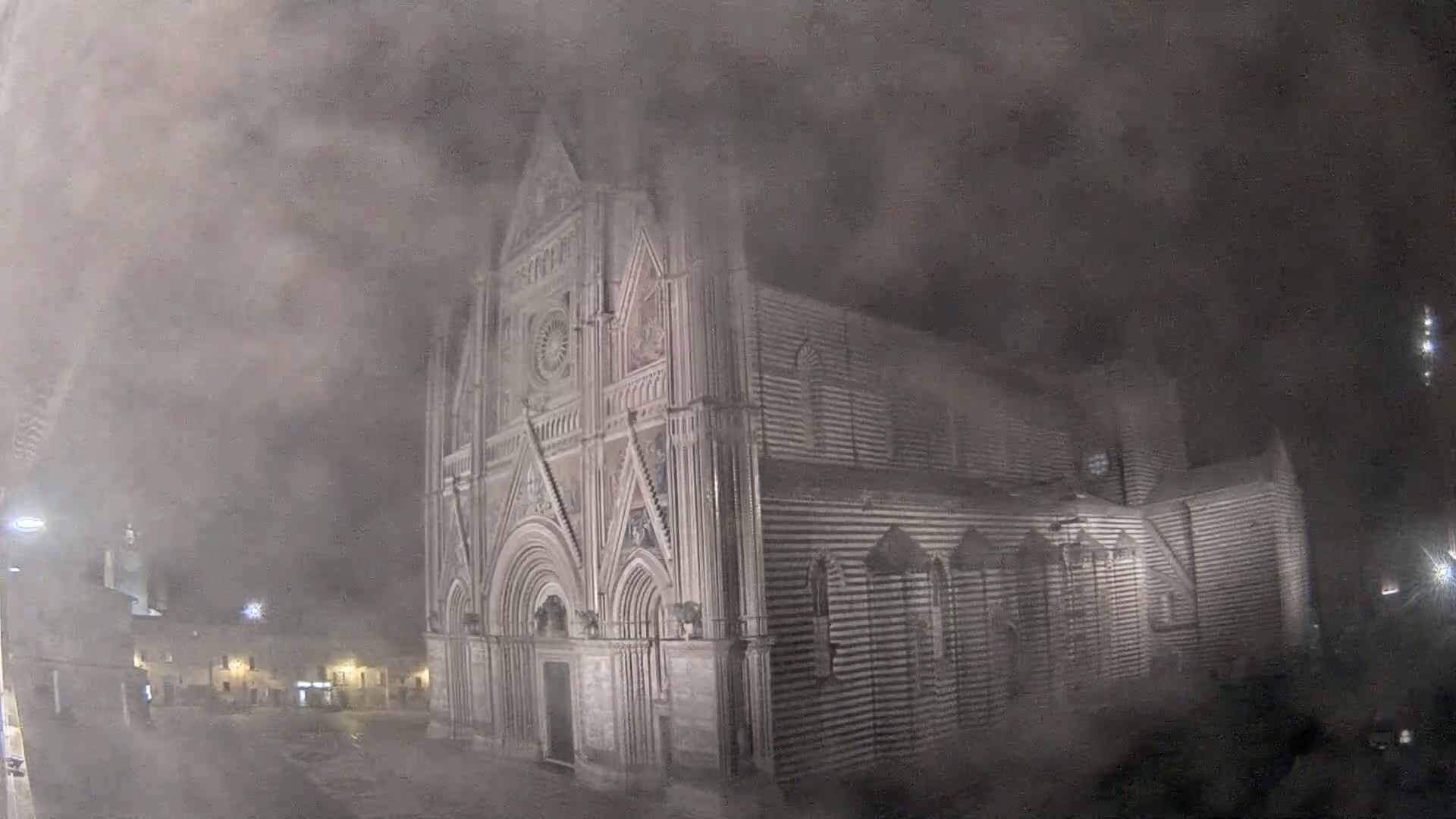 A large, striped stone cathedral stands in a town square at night, shrouded in heavy fog.