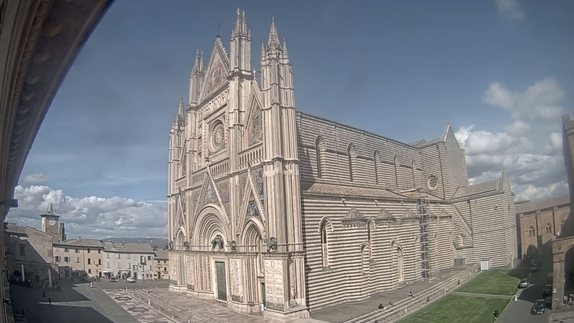 A large, striped cathedral stands in a town square under a mostly sunny sky with some scattered clouds.