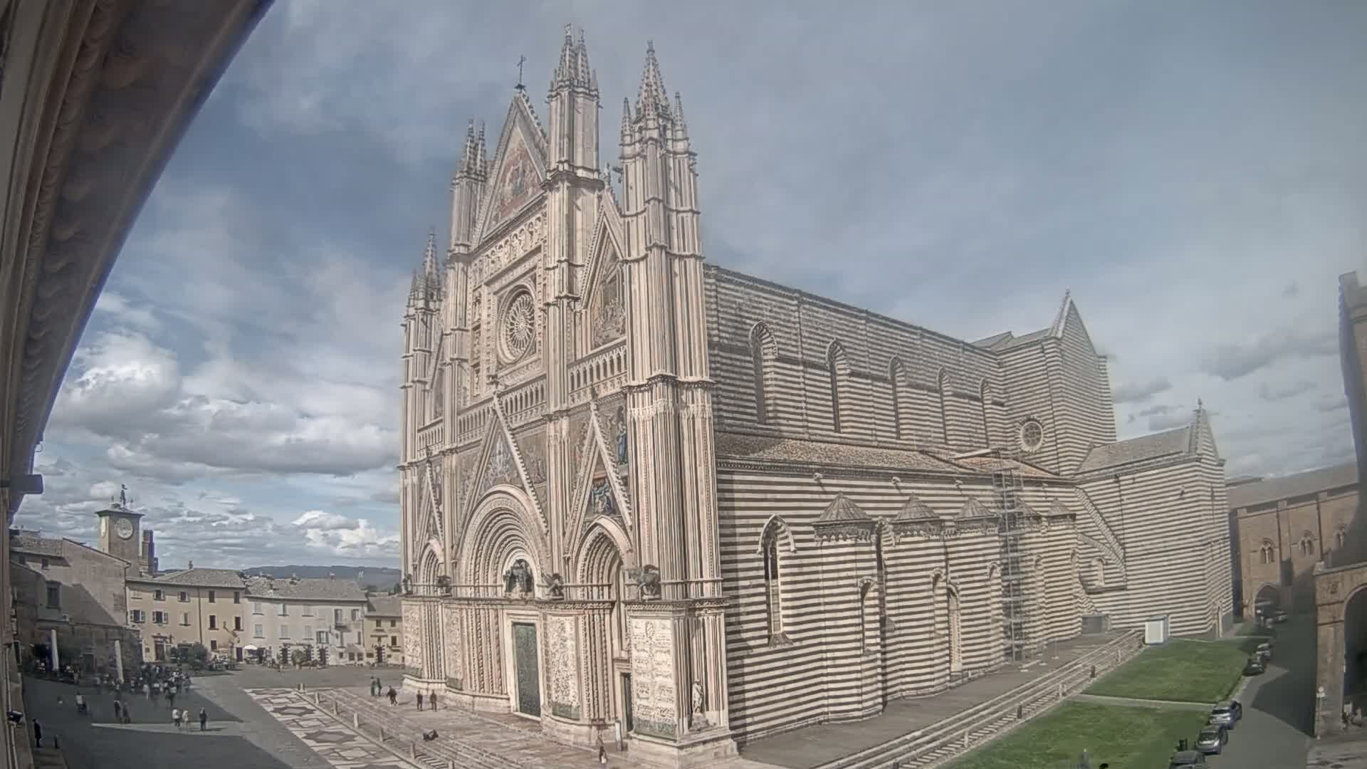A large striped stone cathedral stands in a town square on a partly cloudy day.