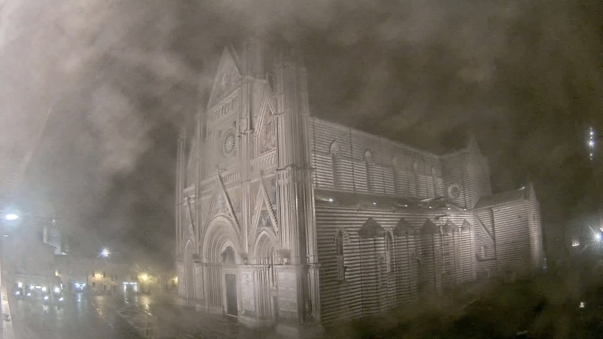 A large, striped stone cathedral is partially obscured by heavy fog or mist at night, with a wet street visible in the foreground.