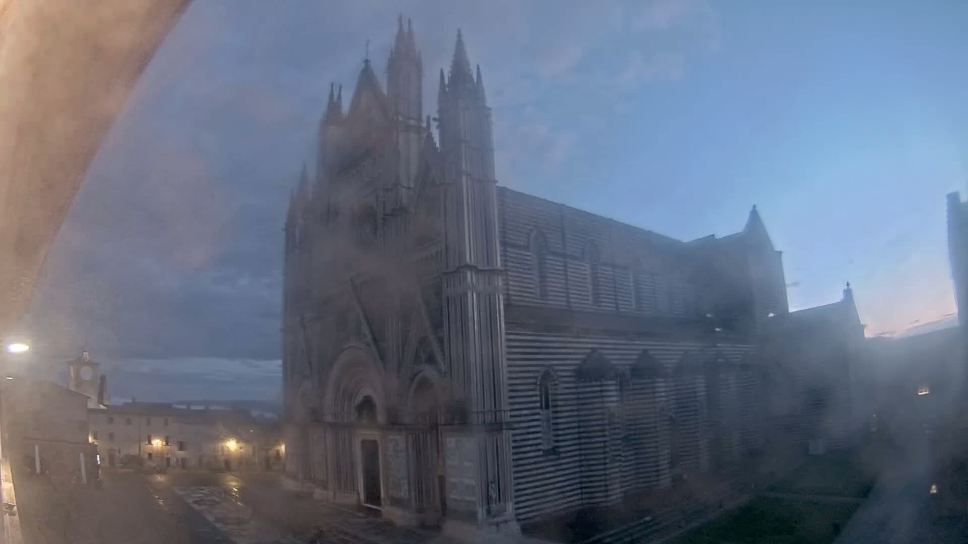A large, striped cathedral stands in a town square on an overcast, possibly rainy day.
