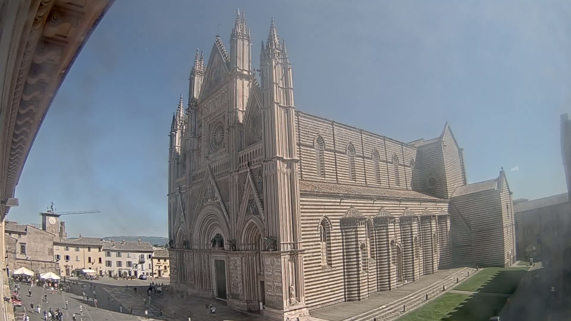 A large striped stone cathedral stands in a sunny town square, with people walking around in front of it.