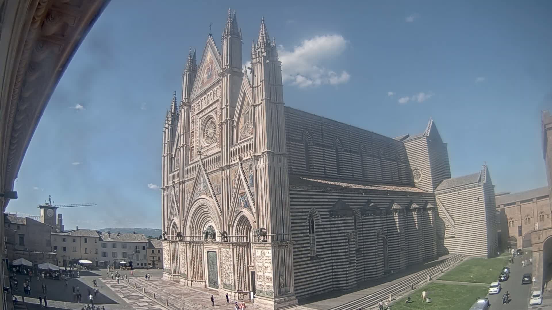 A large, ornate cathedral with striped exterior walls stands in a sunny square with a few people and cars visible in the distance.