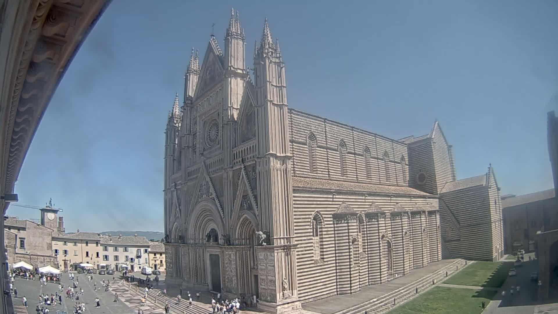 A large striped stone cathedral stands in a sunny town square, surrounded by other buildings and people.