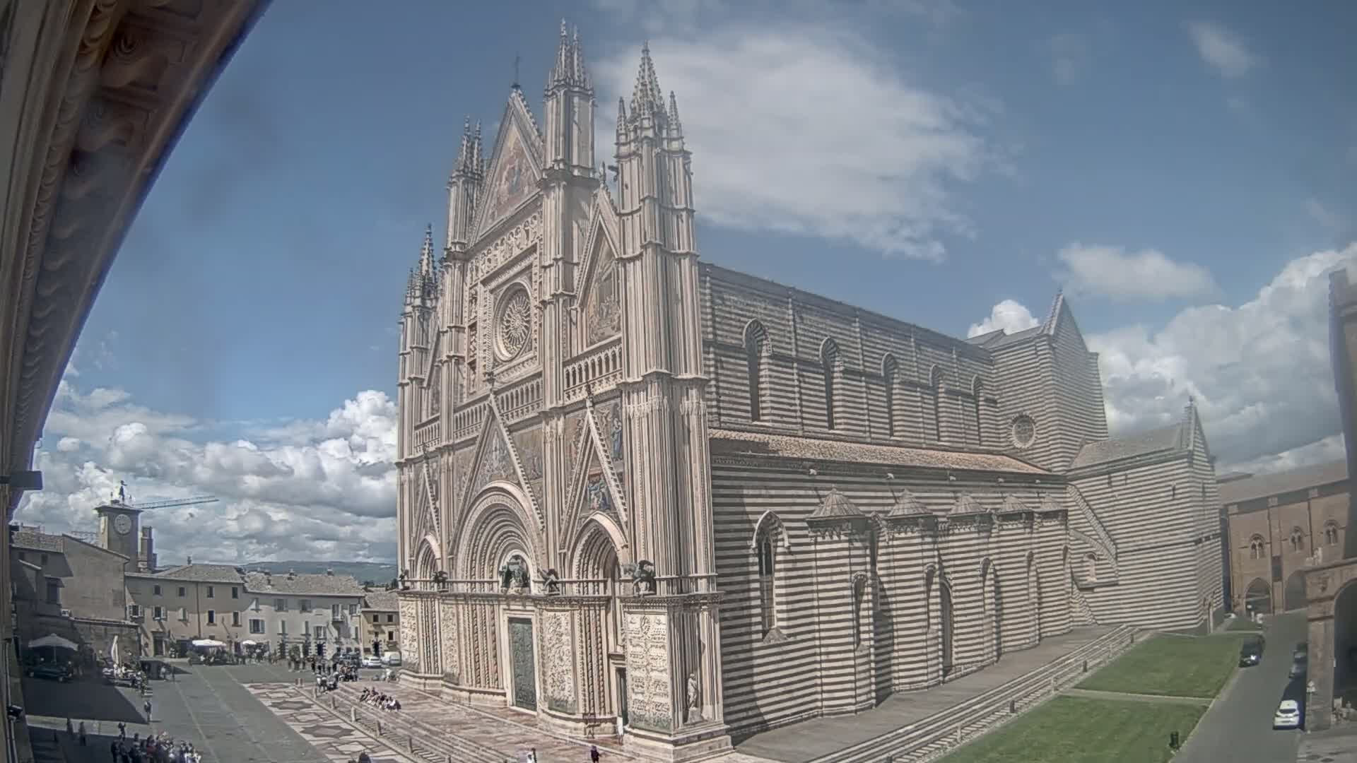A large, ornate cathedral with striped exterior walls stands in a town square under a partly cloudy sky.