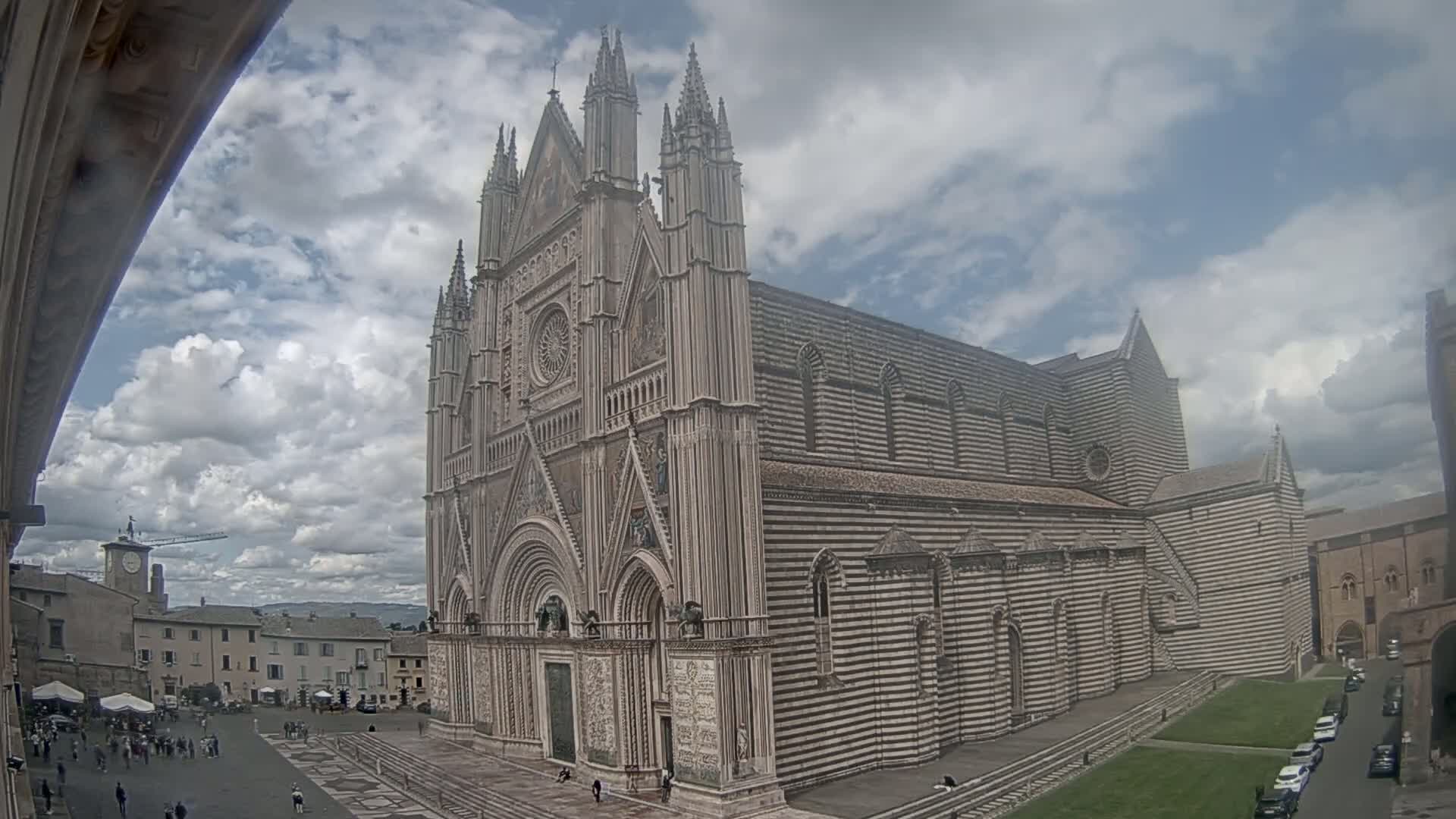 A large, striped cathedral with pointed spires stands in a town square under a partly cloudy sky.