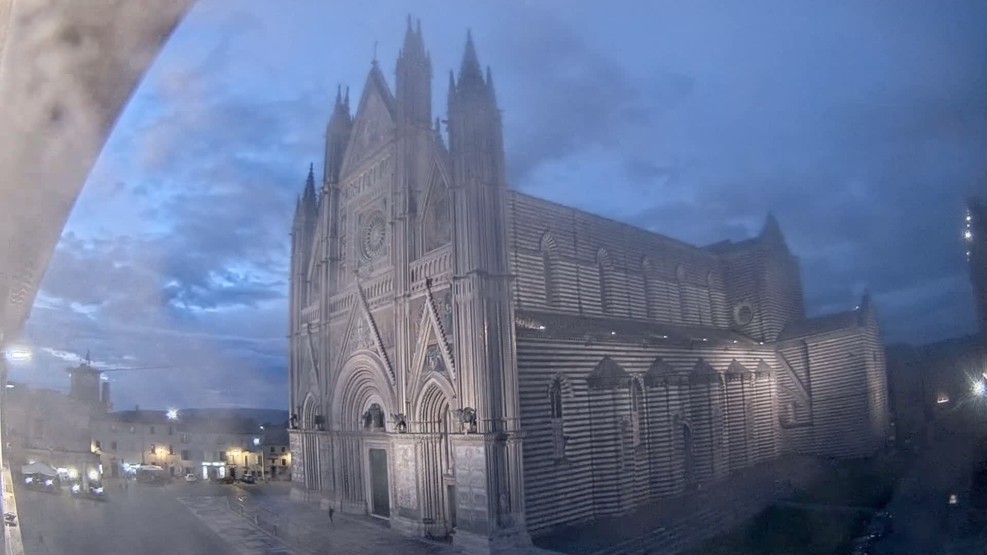 A large, striped Gothic cathedral stands in a town square at dusk under a cloudy sky.