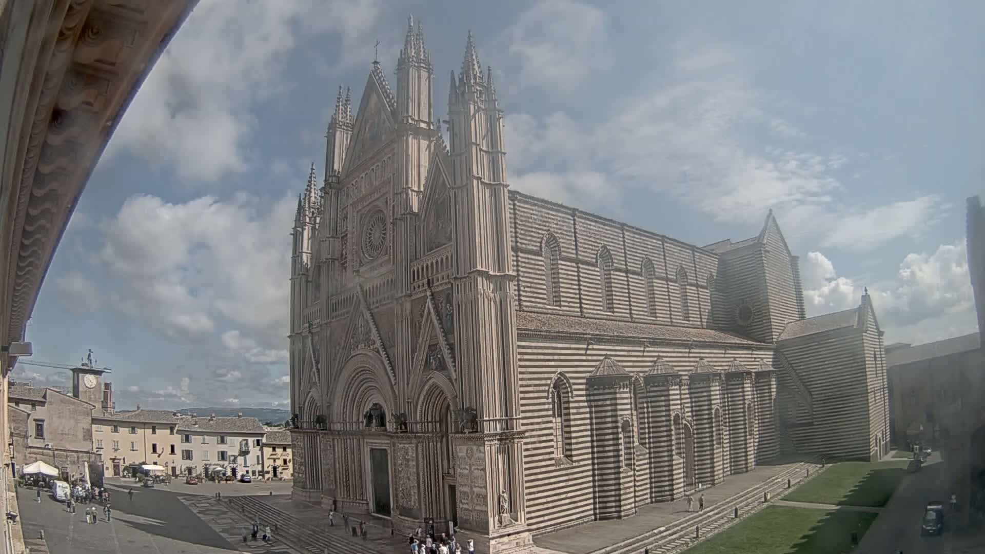 A large, striped cathedral stands in a town square under a partly cloudy sky.