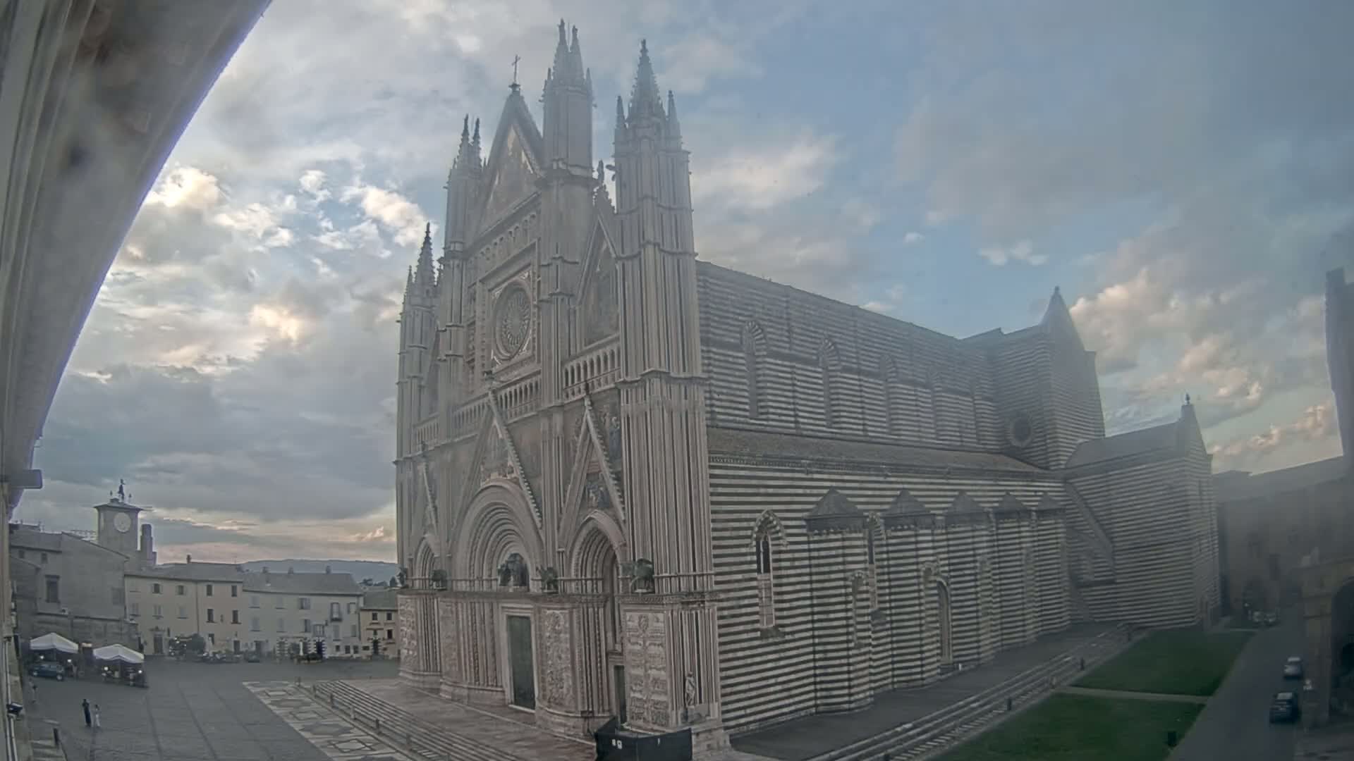 A large, striped cathedral stands in a town square under a partly cloudy sky.