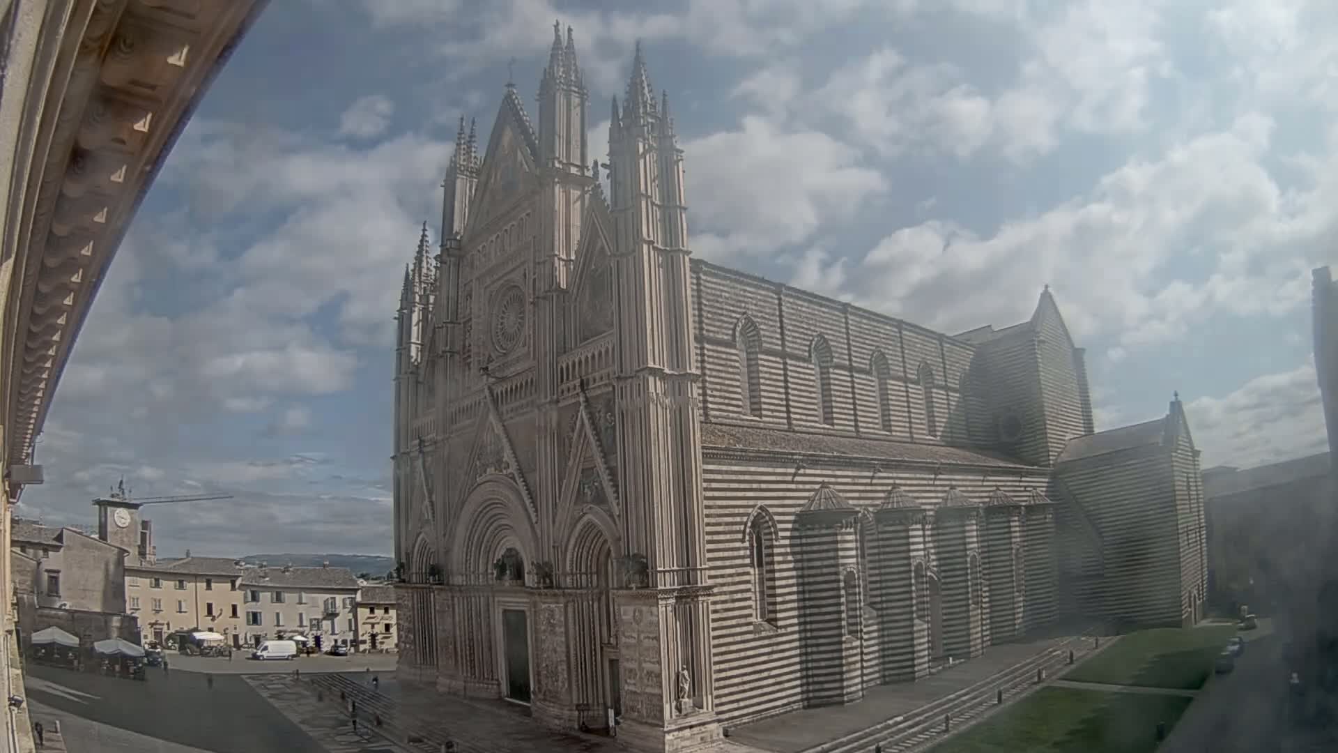A large, striped stone cathedral stands in a town square under a partly cloudy sky.