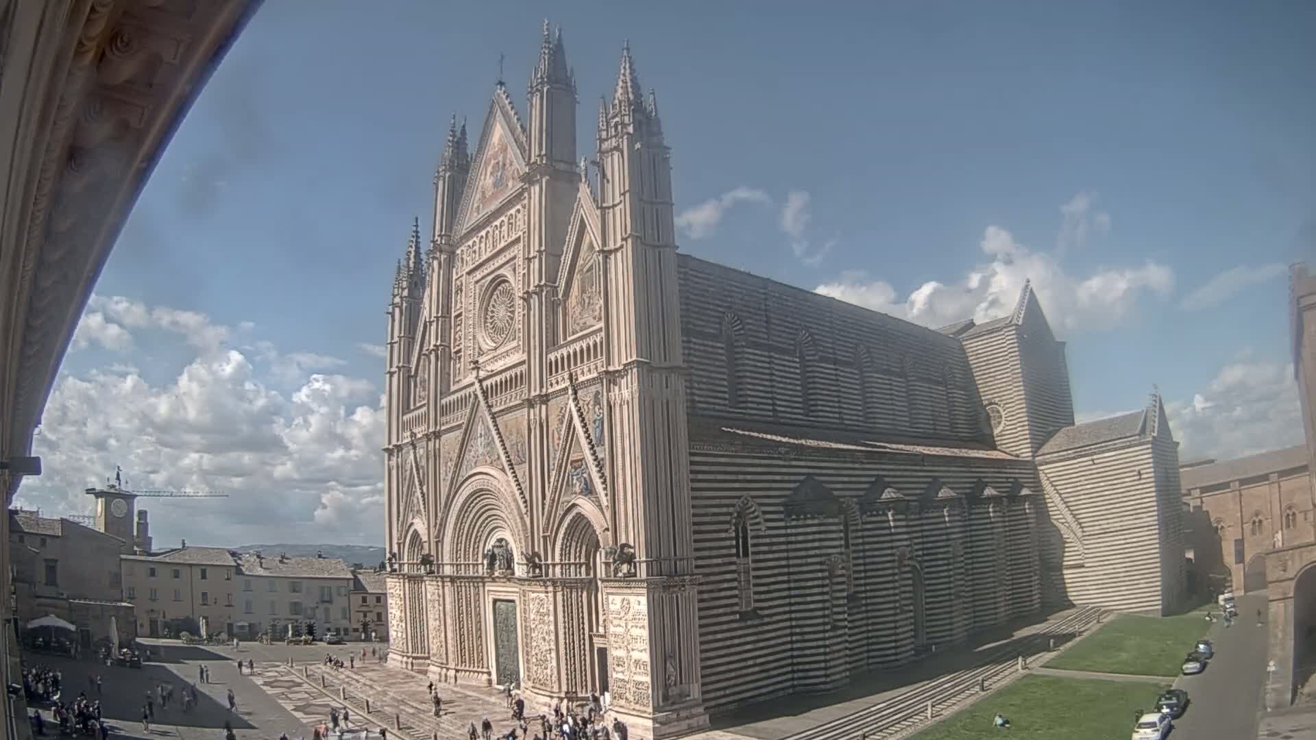 A large, striped cathedral stands in a town square under a partly cloudy sky.