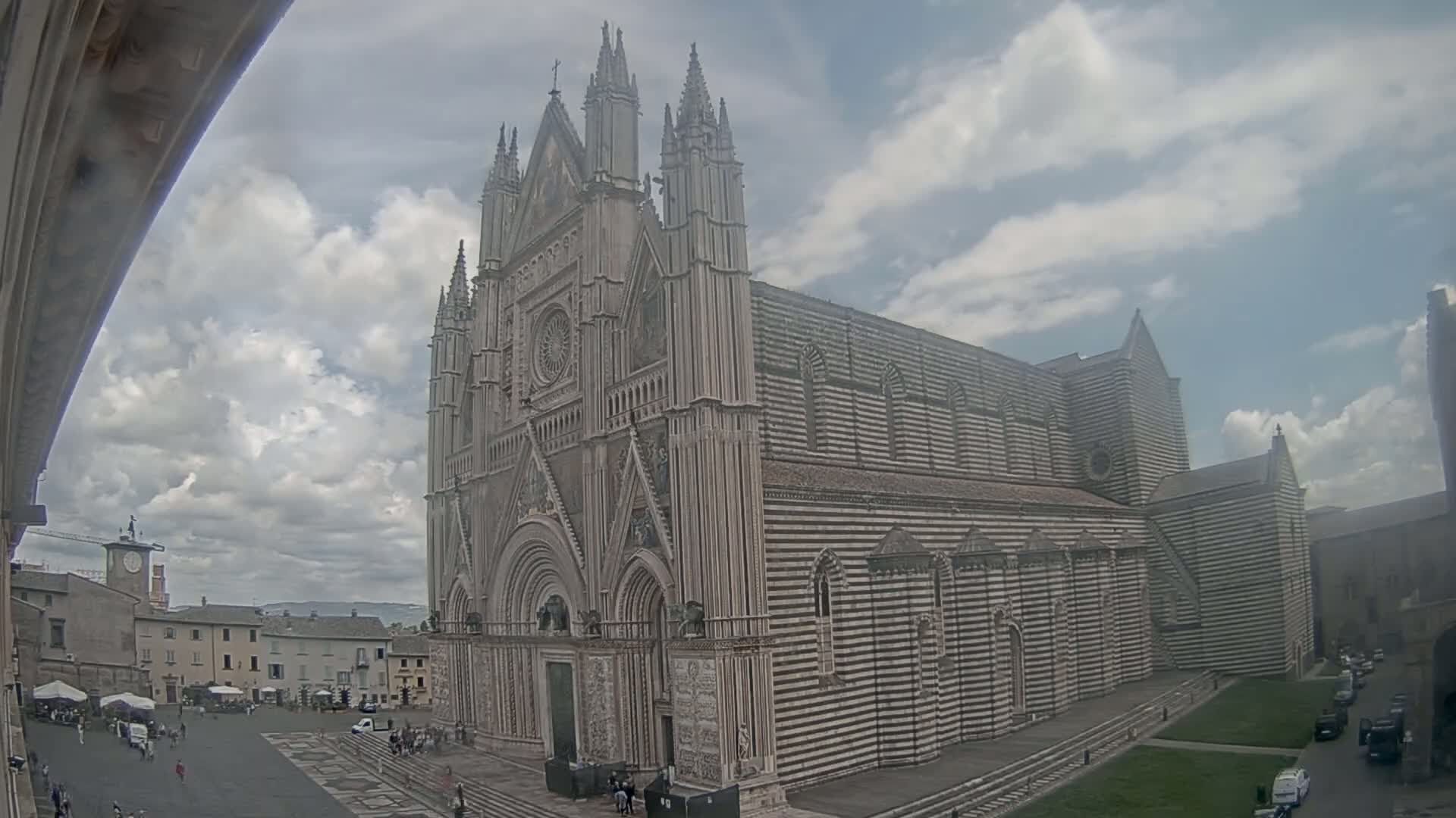 A large, striped cathedral stands in a town square under a partly cloudy sky.