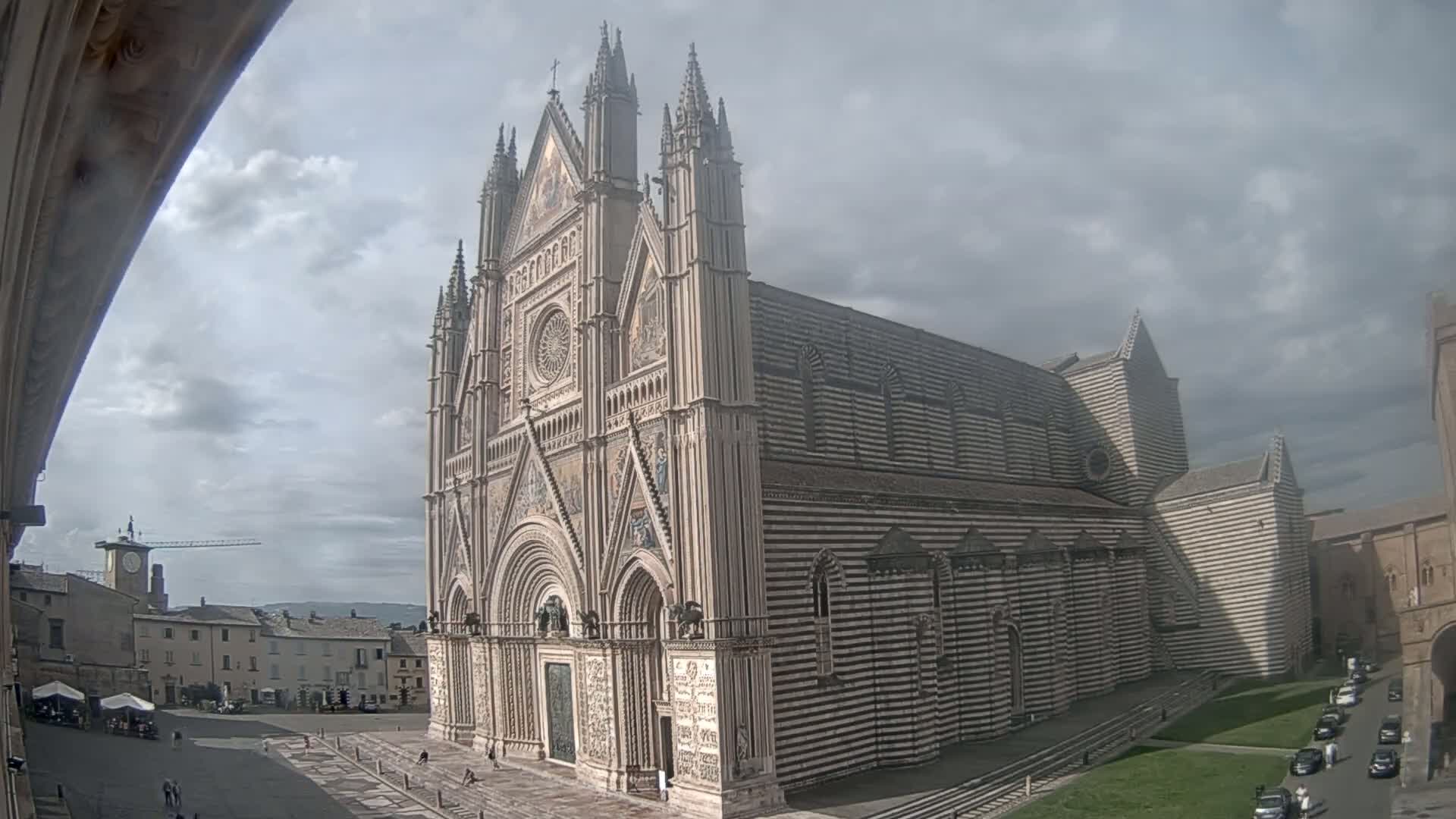 A large cathedral with striped exterior walls stands in a town square under a cloudy sky.