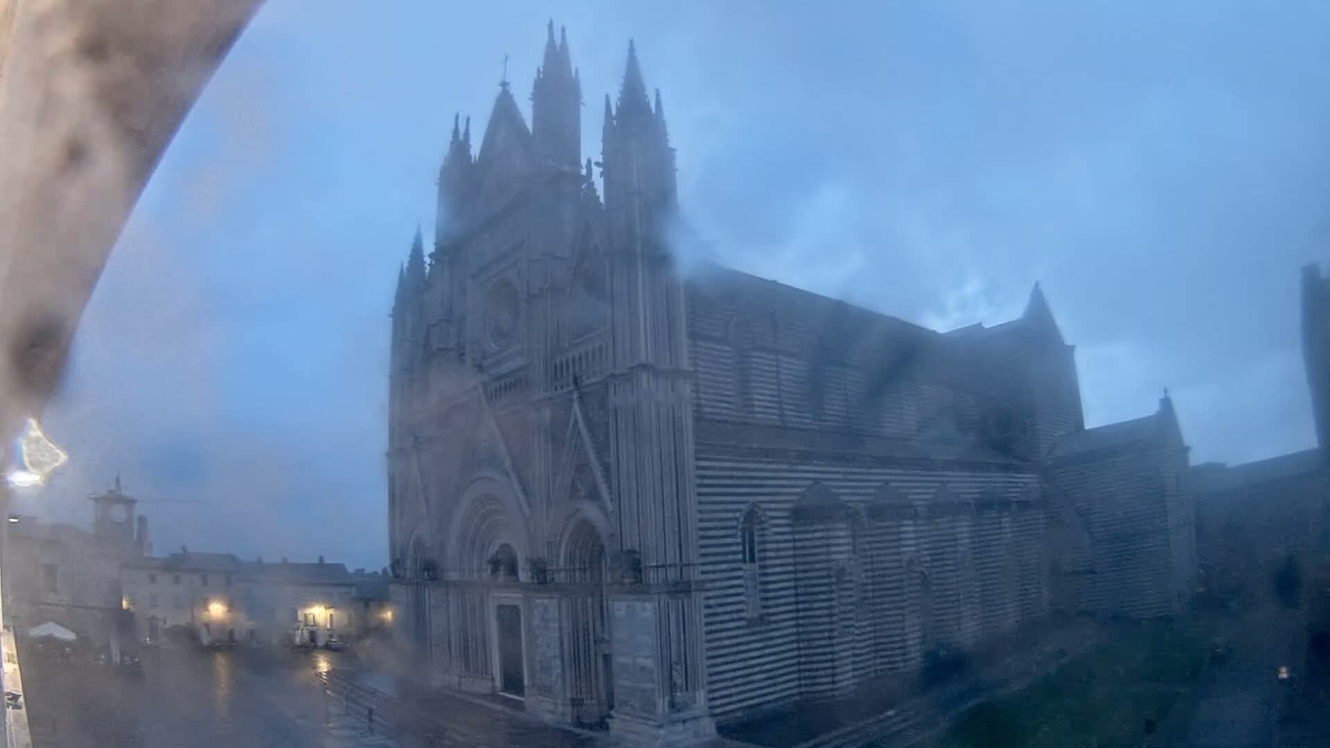 A large gothic cathedral with striped exterior walls is seen through rain-streaked glass on a gloomy day.
