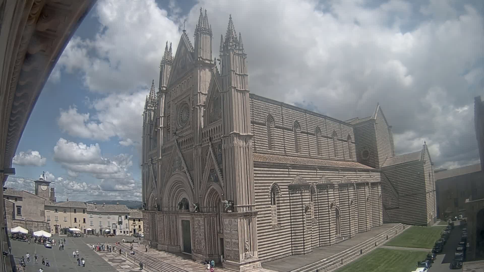 A large, striped stone cathedral stands in a town square under a partly cloudy sky.