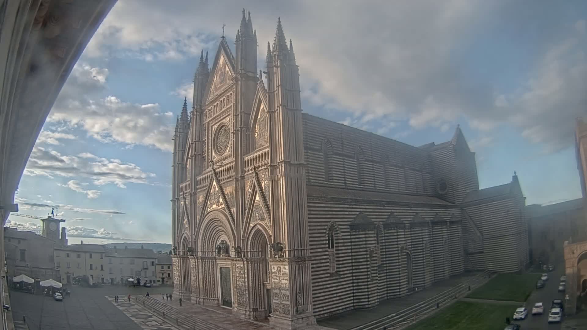 A large, striped cathedral stands in a town square under a partly cloudy sky.