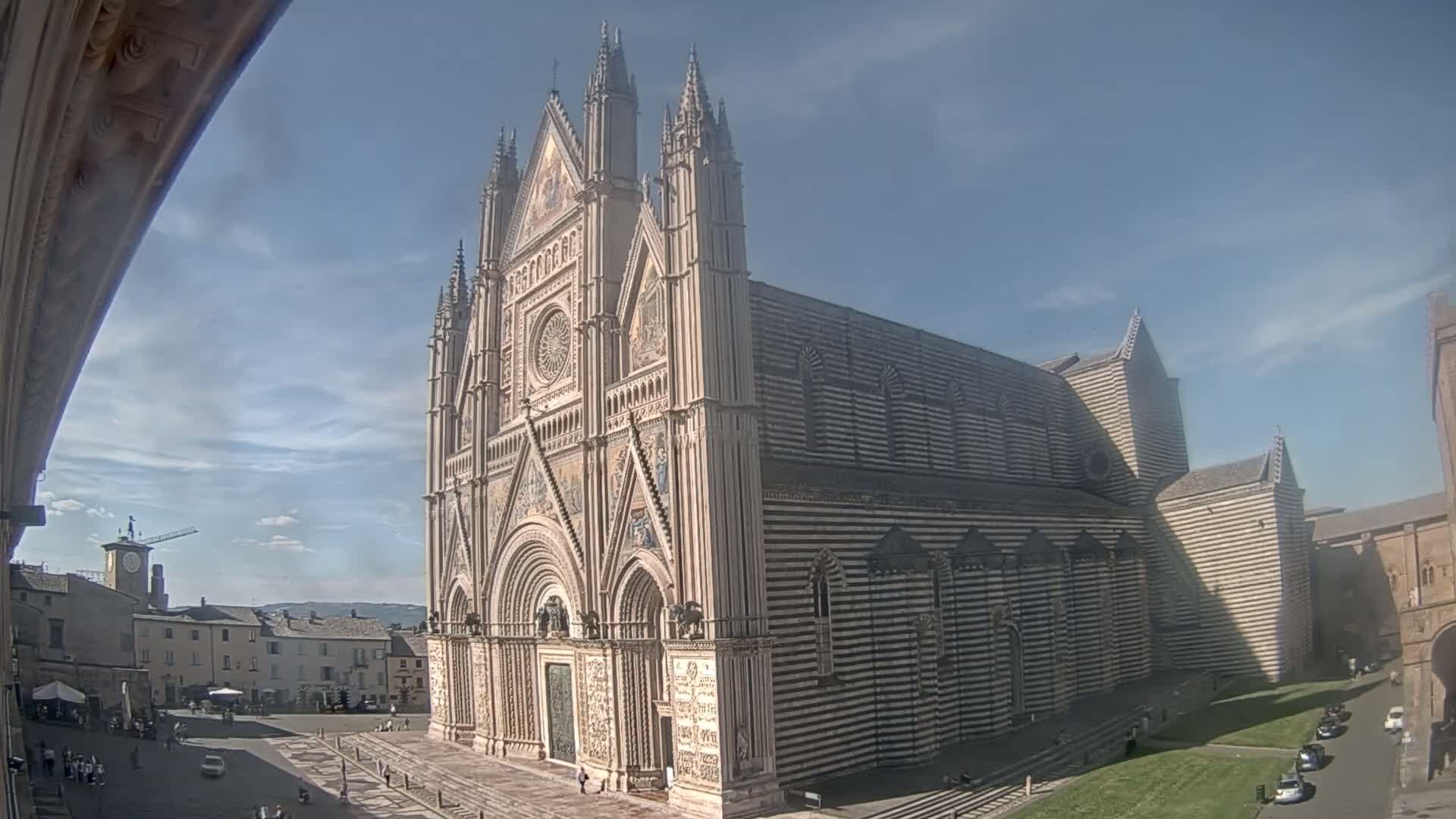 A large, striped cathedral stands in a sunny square with a few people and cars visible, under a mostly clear sky.