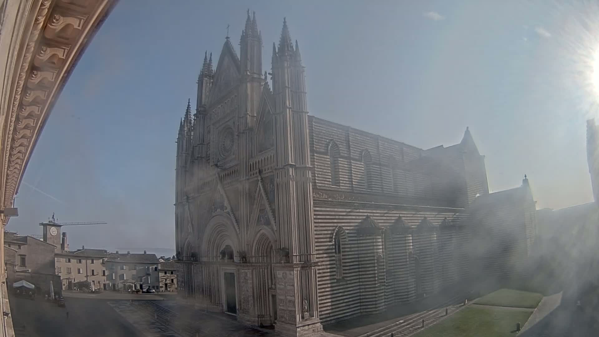 A large, striped stone cathedral stands in a hazy, sunny outdoor setting, with a town square and buildings visible in the background.