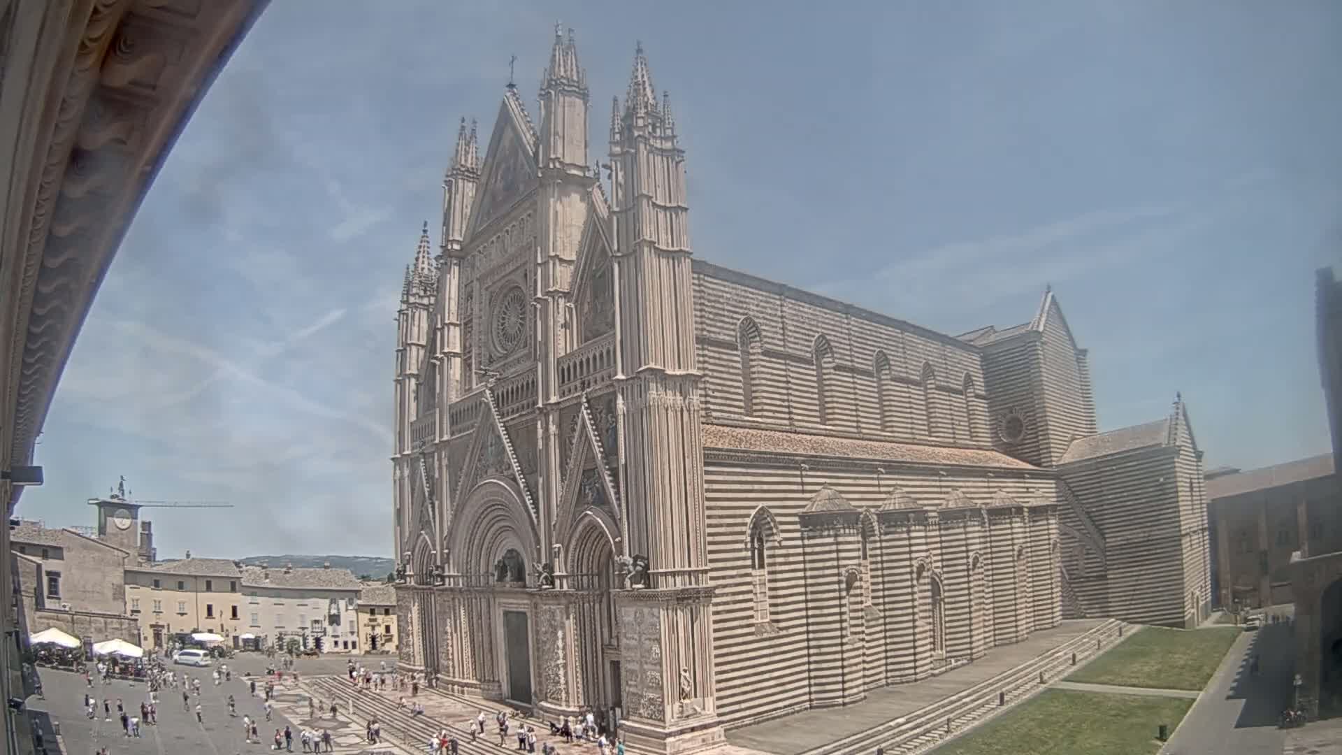 A large, ornate cathedral with striped stonework stands in a sunny plaza surrounded by buildings and people.