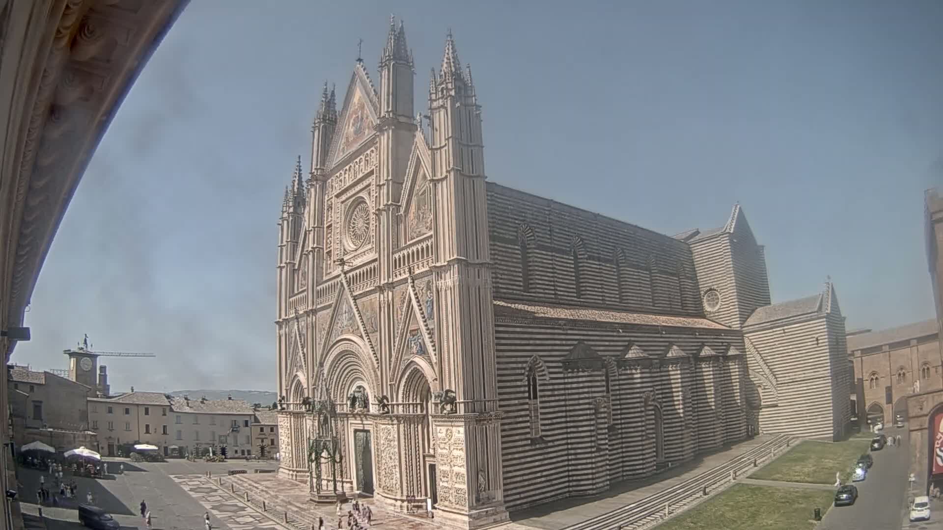 A large, striped cathedral stands in a sunny, city square with a few people and cars visible.