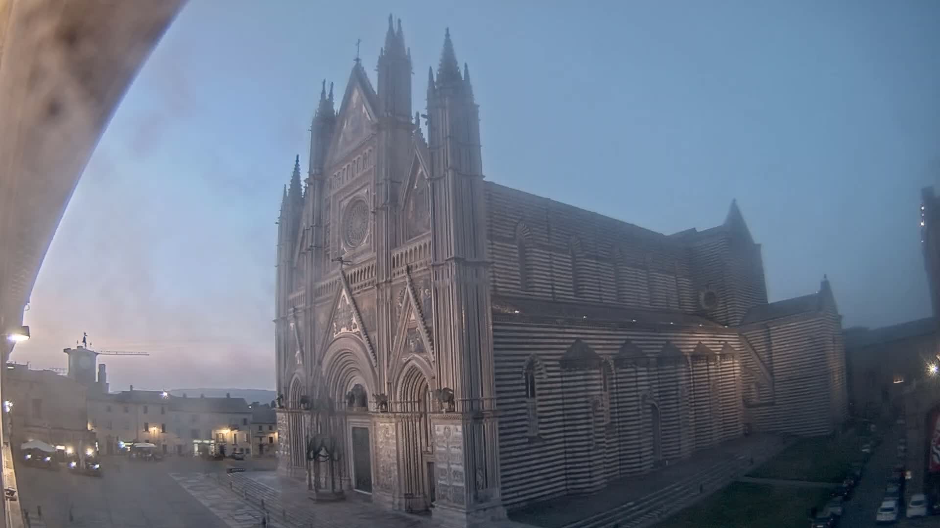 A large, striped cathedral stands in a town square shrouded in early morning fog.