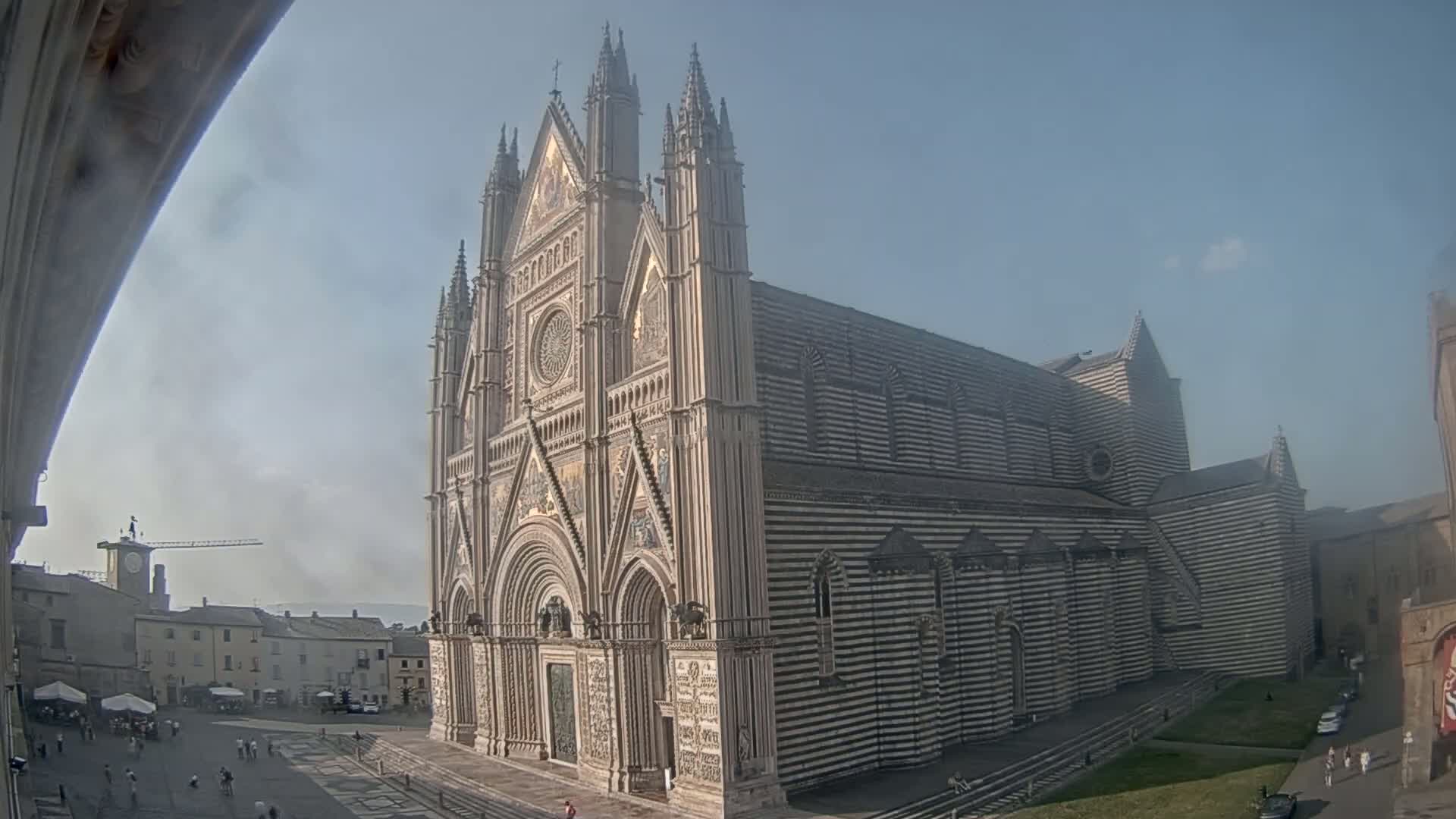 A large, striped cathedral with a Gothic facade stands in a town square on a hazy, sunny day.