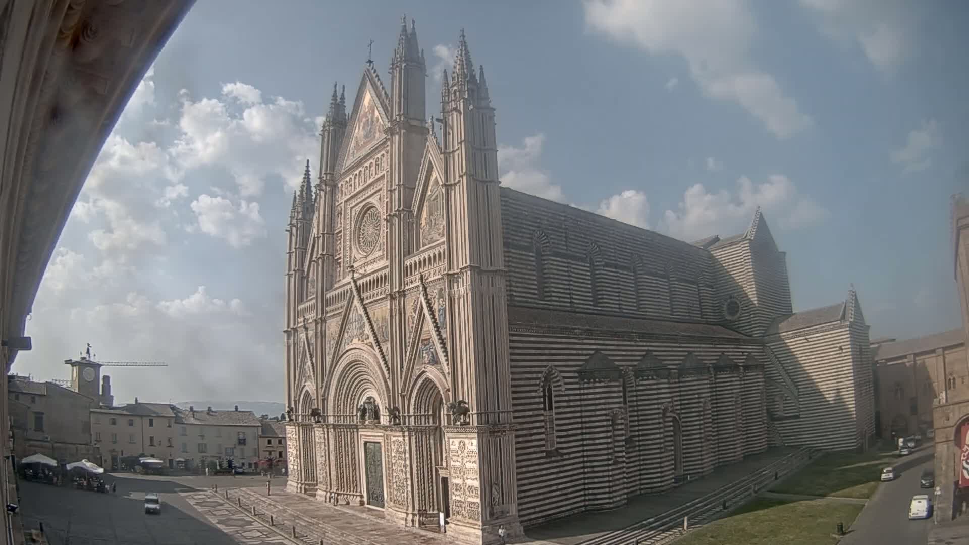 A large, striped cathedral stands in a town square under a partly cloudy sky.
