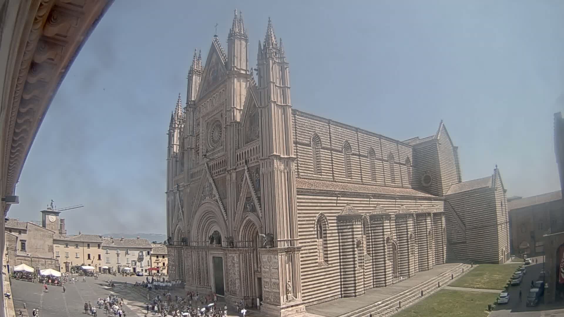 A large striped stone cathedral stands in a town square on a sunny day, with people gathered in the square below.