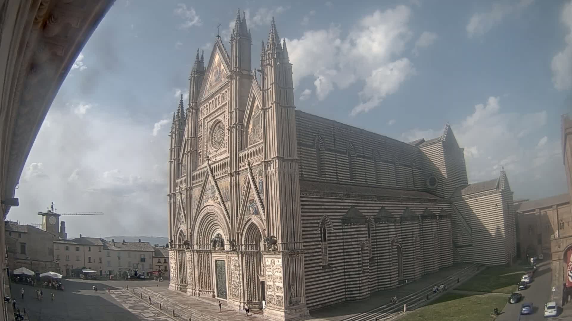 A large, striped Gothic cathedral stands in a town square under a partly cloudy sky.