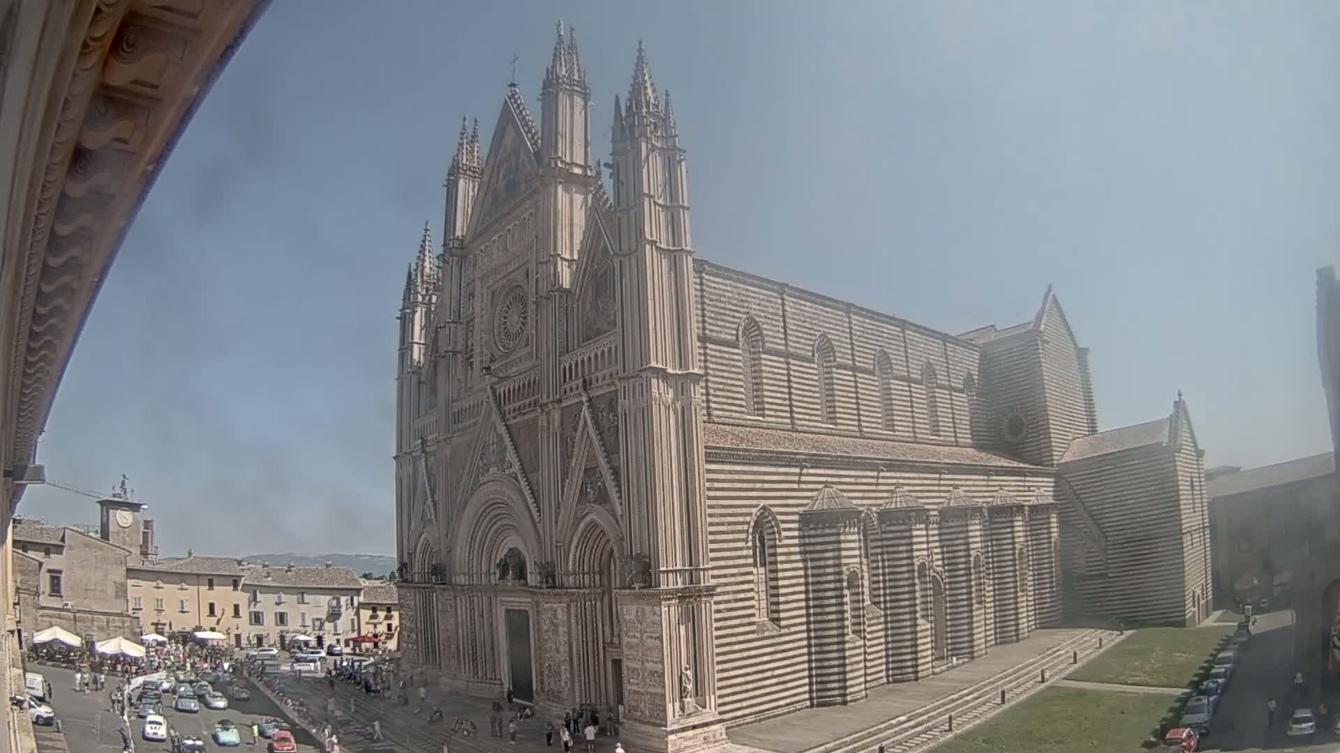 A large, striped stone cathedral stands prominently in a town square on a sunny day, with cars and people visible in the foreground.
