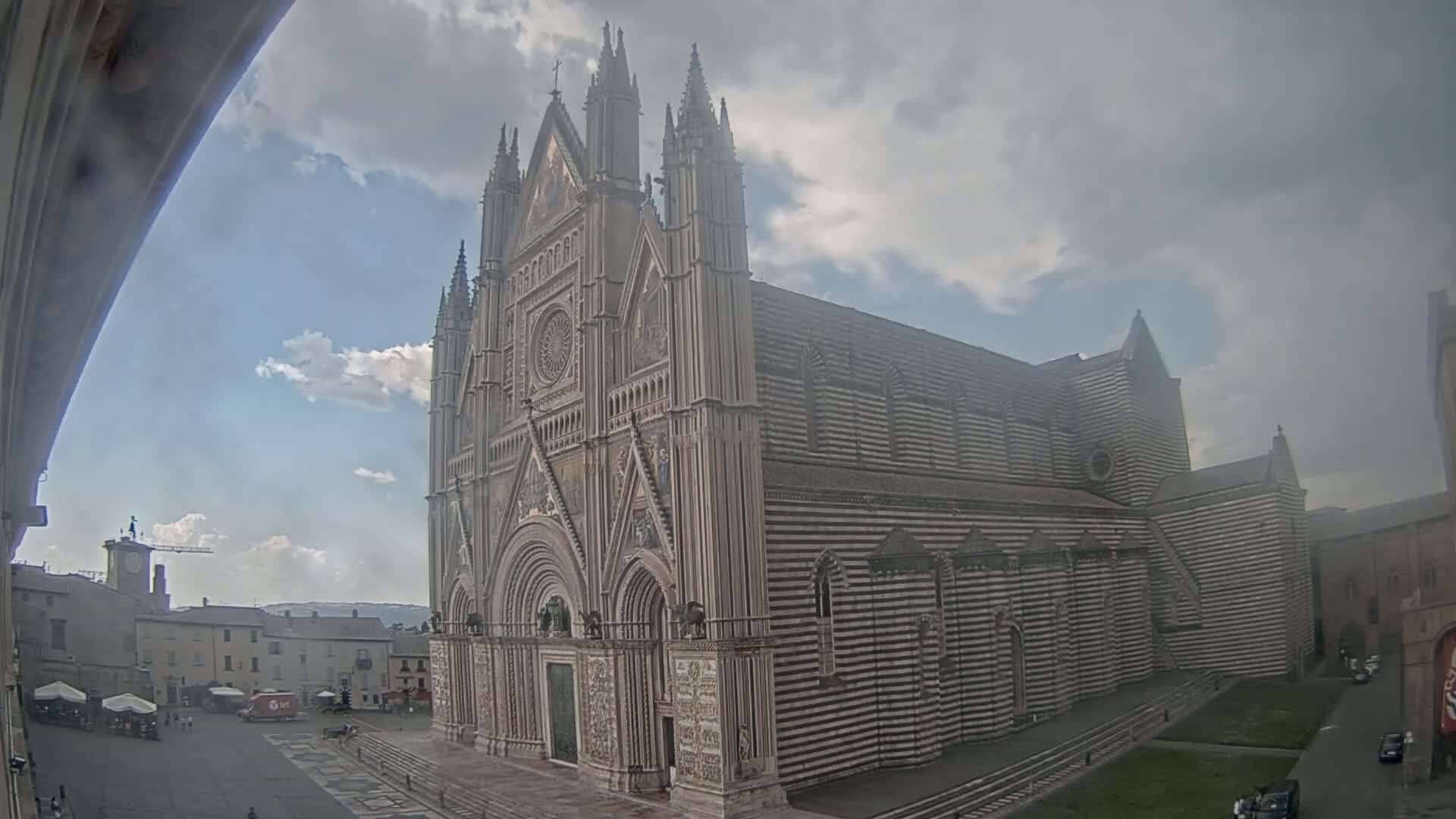 A large, striped cathedral with Gothic architectural details stands in a town square under a partly cloudy sky.