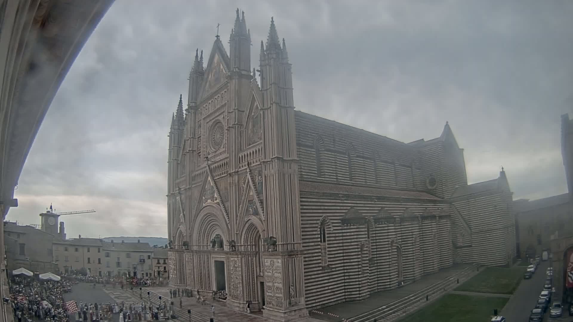 A large, striped cathedral stands in a town square under an overcast sky, with a crowd of people gathered in the foreground.