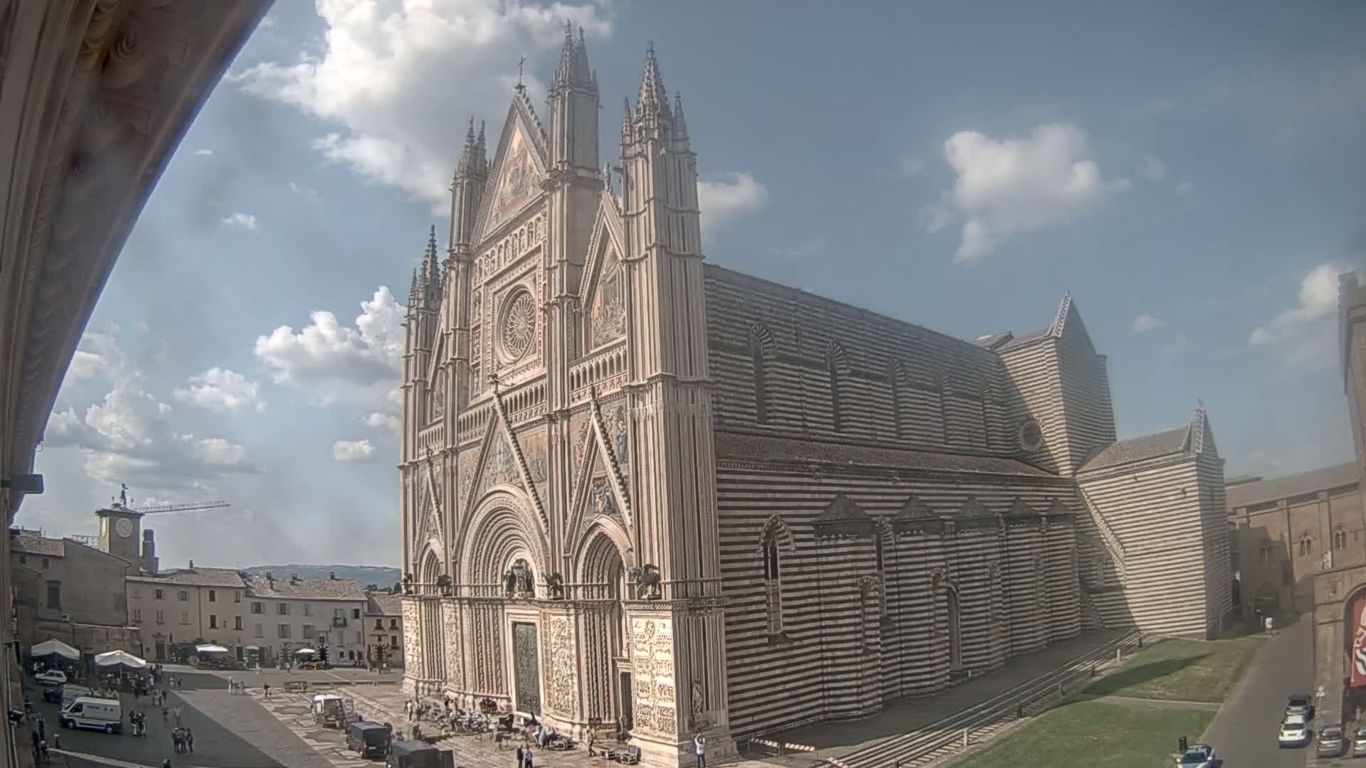 A large, striped cathedral stands in a town square under a partly cloudy sky.