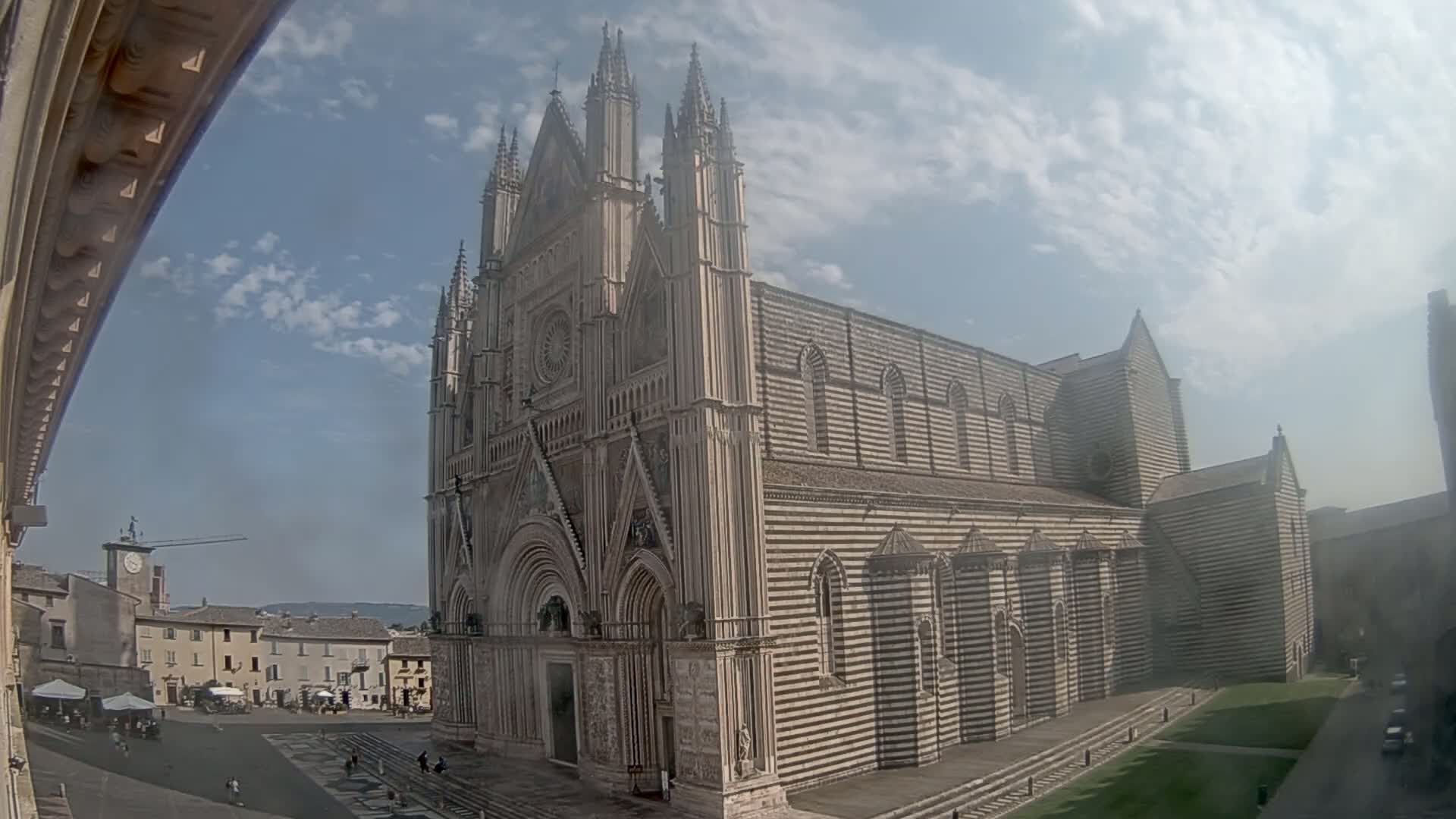 A large, striped, Gothic-style cathedral stands in a town square under a partly cloudy sky.