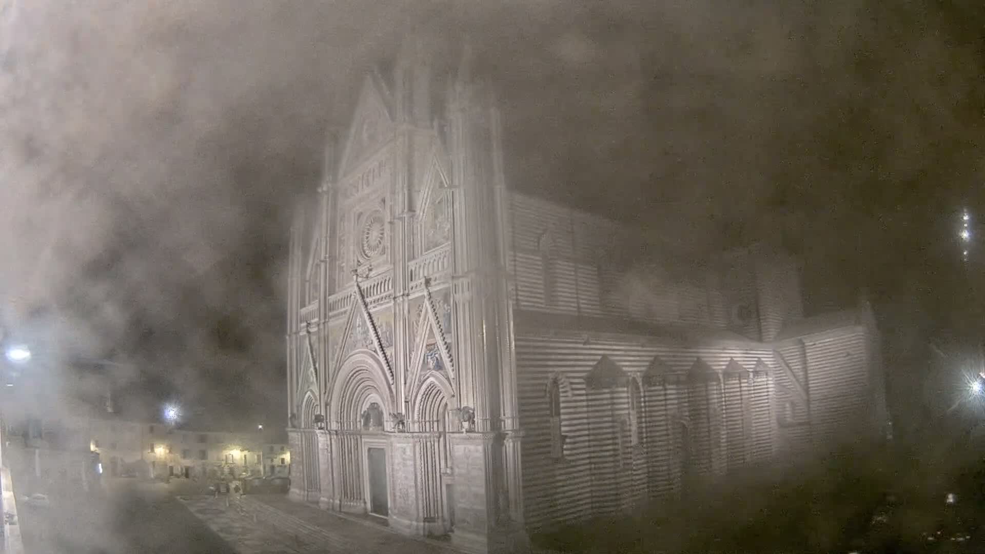 A grand, intricately detailed cathedral with a striped side stands illuminated at night, largely enveloped by dense fog that also obscures the adjacent city square.