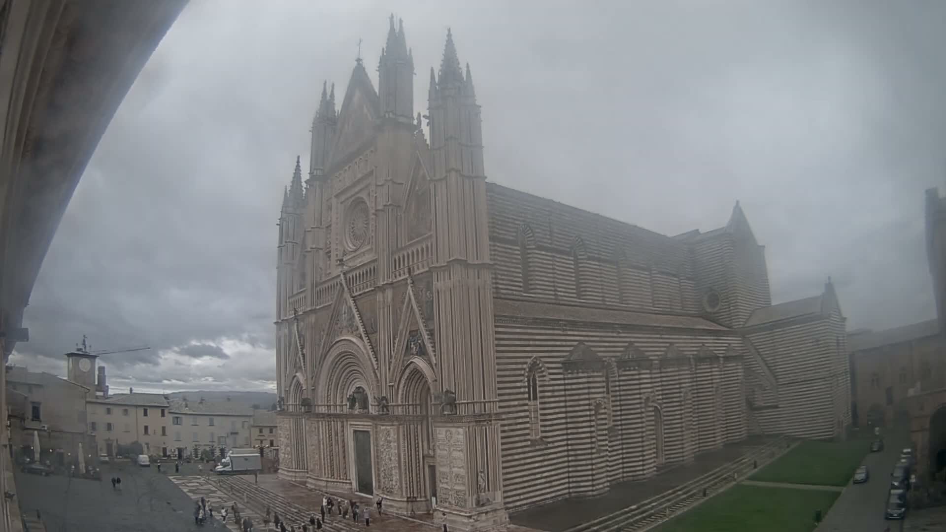 A grand, intricately detailed cathedral with a striped side stands illuminated at night, largely enveloped by dense fog that also obscures the adjacent city square.