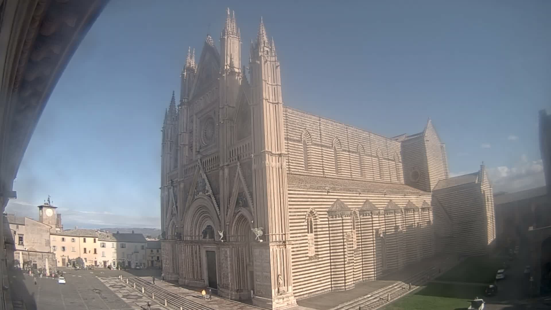 A grand, striped marble cathedral with multiple spires stands prominently beside a town square featuring a clock tower and other buildings, all under a clear blue sky on a bright day.