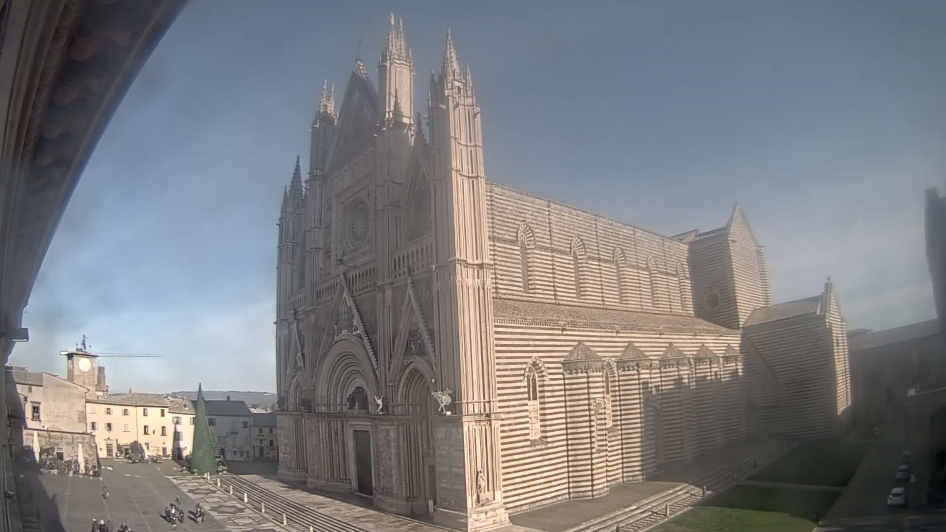 A grand, ornate striped cathedral stands prominently overlooking an outdoor city square bustling with people, all under a clear blue, sunny sky.