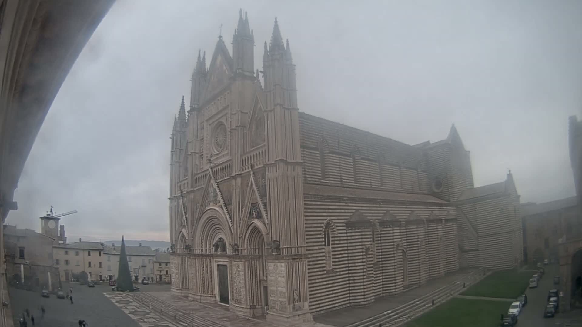 A grand, ornate Gothic cathedral with a distinctive horizontally striped facade dominates a misty town square, flanked by a clock tower, other buildings, and a large conical structure, all under a heavily overcast sky.