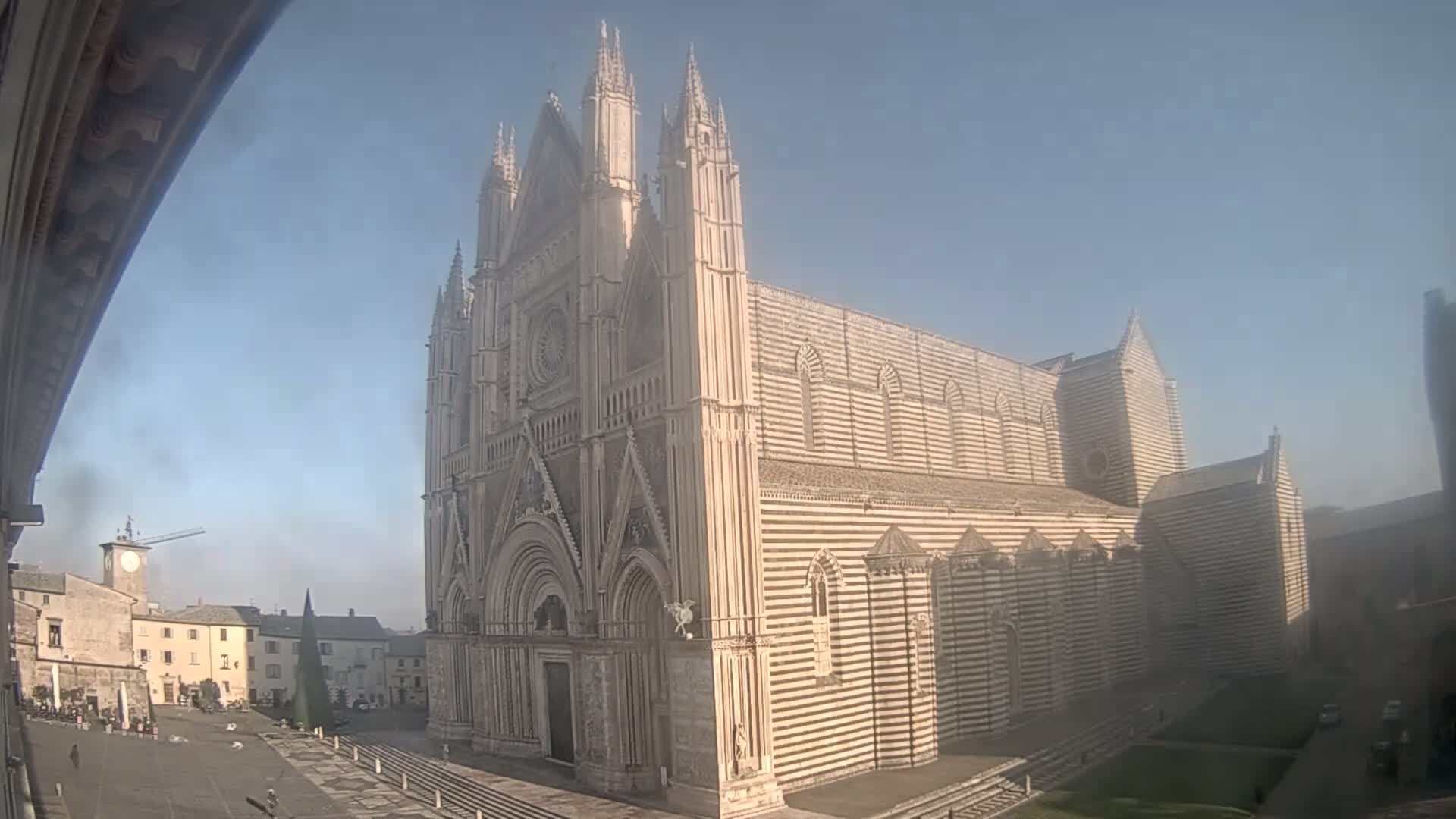 A grand, ornate Gothic cathedral with a distinctive horizontally striped facade dominates a misty town square, flanked by a clock tower, other buildings, and a large conical structure, all under a heavily overcast sky.