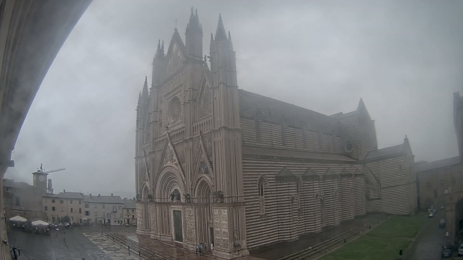 A grand, ornate Gothic cathedral with a distinctive horizontally striped facade dominates a misty town square, flanked by a clock tower, other buildings, and a large conical structure, all under a heavily overcast sky.