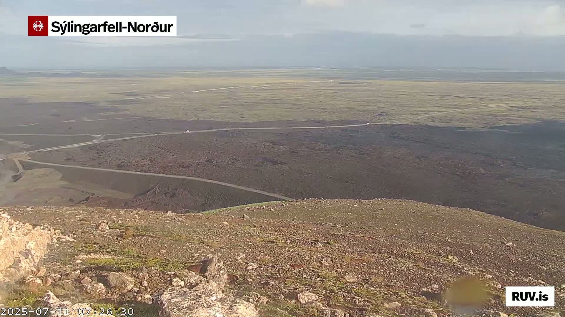 A high-angle view shows a rocky, sparsely vegetated landscape with winding roads leading to a flat, grassy plain under a mostly clear sky.