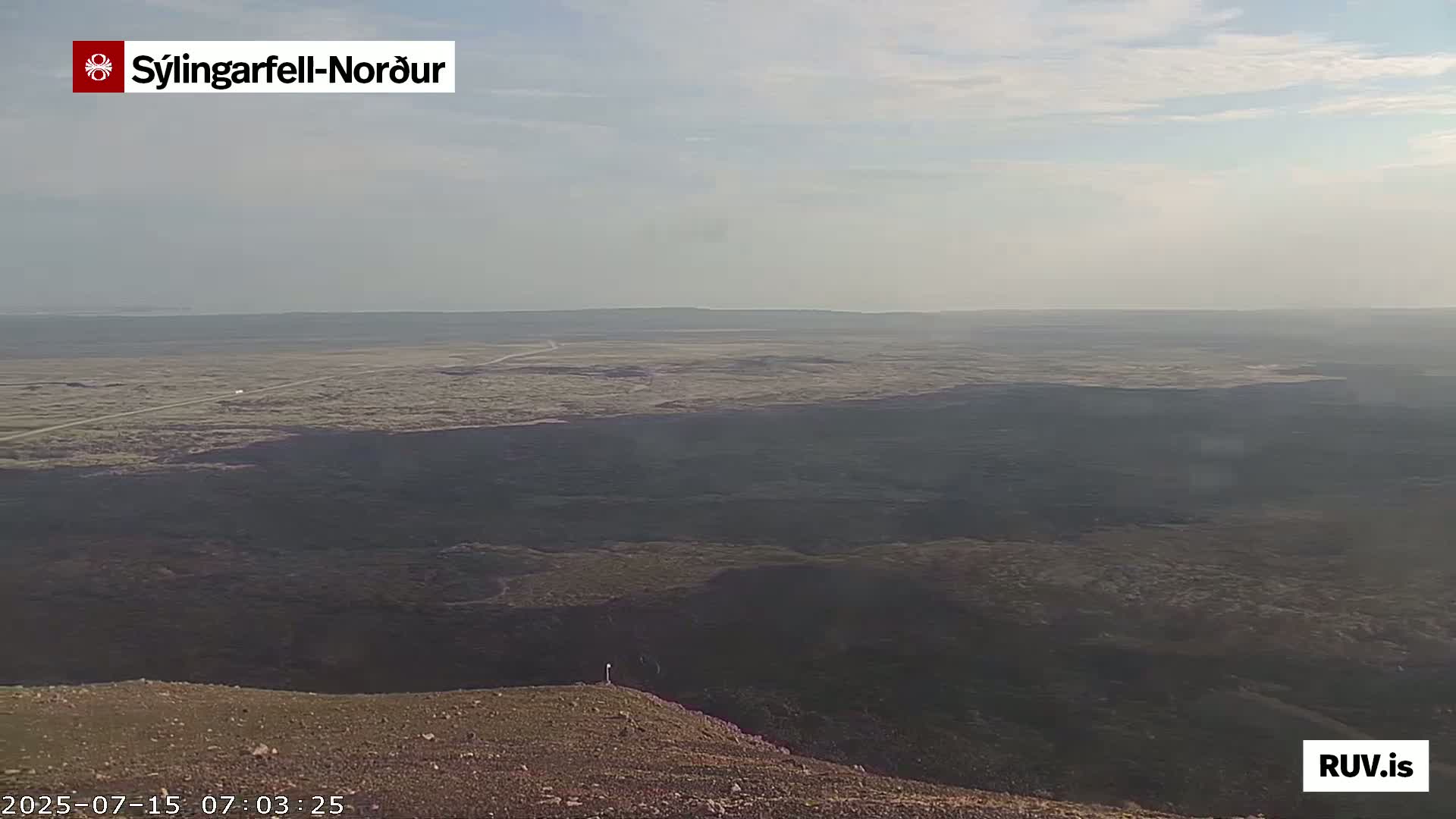 A vast, flat, dark brown landscape stretches to a hazy horizon under a mostly clear sky.