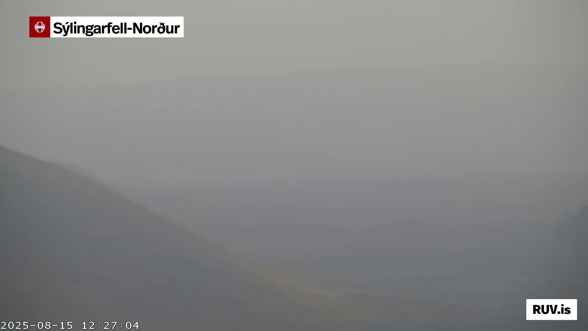 A hazy, long shot shows a vast, dark, mountainous landscape under a pale sky, with wisps of smoke or steam visible in the distance.