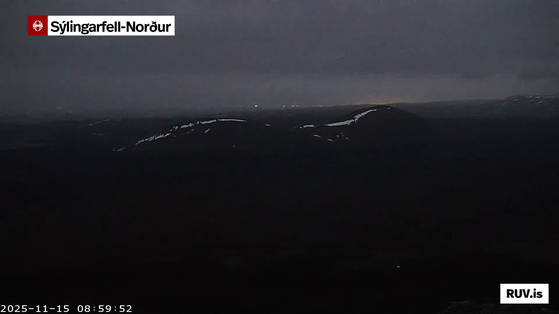 A dark, wide shot of a mountainous landscape at night or very early morning under a heavily overcast sky, with distant faint lights of a settlement and patches of snow visible on the ridges.